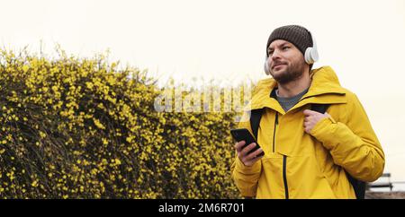Adult man in yellow jacket in hat and headphones holding mobile phone in hand against plants with yellow flowers. Stock Photo
