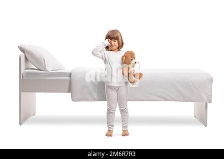 Girl in pajamas holding a teddy bear and rubbing eyes in front of a bed isolated on white background Stock Photo