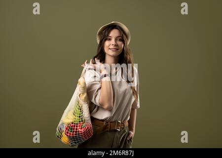 Portrait of brunette woman holding cotton net shopper, reusable mesh shopping bags with vegetables, products on pastel green background. Eco friendly Stock Photo