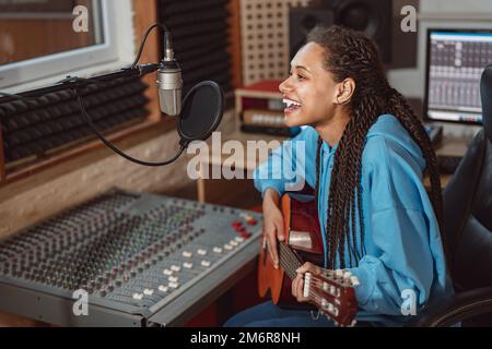 Enthusiastic African young woman musician plays guitar and records song, sings into microphone in music recording studio Stock Photo