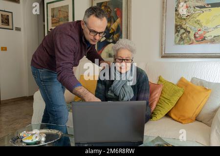Man helping an old woman with the laptop. Woman sitting on the couch in her living room with a laptop on the table in front of her. Stock Photo