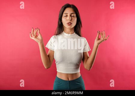 Young caucasian woman wearing casual top isolated over red background keeping eyes closed, exhales, holding fingers in mudra gesture. Meditation, reli Stock Photo