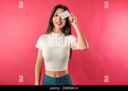Young dark-haired woman wearing ribbed crop isolated over red background holds credit card over one eye and poses for the camera. Stock Photo