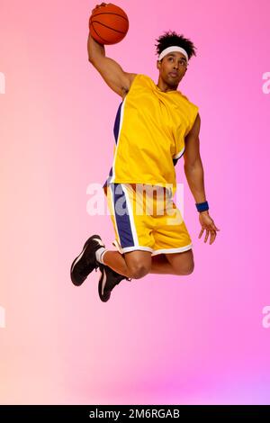 Image of biracial basketball player in the air with basketball on pink to orange background Stock Photo