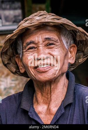 Vertical portrait of South Asian Balinese senior man wearing a traditional Balinese cone-shaped hat. An elderly man looks into the camera smiling. Stock Photo