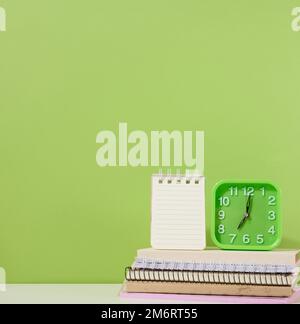 An open notepad and a green alarm clock stands on a stack of notebooks, a green background. Stock Photo