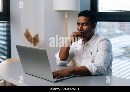 Pensive young African-American businessman working on business strategy thinking looking to laptop screen sitting at desk in modern office room. Stock Photo