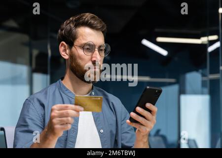 Serious thinking man inside office holding bank credit card and smartphone, businessman making money transfer, reservation and purchase in online store, successful entrepreneur. Stock Photo