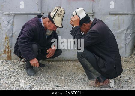 Murghab, Gorno-Badakshan, Tajikistan - 07 30 2014 : Two old men wearing traditional kyrgyz kalpak felt hat squatting and talking in Murghab market Stock Photo