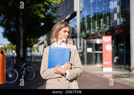Work and corporate people. Confident businesswoman holding a blue folder with business documents, walking on street Stock Photo