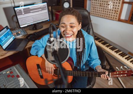 Stylish African pretty woman playing guitar and singing into microphone during rehearsal for concert in recording studio Stock Photo
