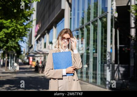 Portrait of corporate woman in sunglasses and beige suit, holding blue folder with office documents, going to work, walking on s Stock Photo