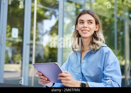Young digital nomad, woman with tablet, working outdoors on fresh air, smiling and looking away at passerby Stock Photo