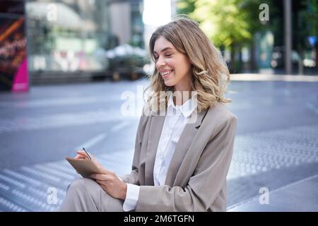 Young businesswoman preparing for speech, interview in company, writing down notes while sitting outside in city centre Stock Photo