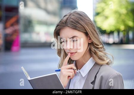 Woman looking concentrated while reading her notes, preparing for interview. Businesswoman in suit writes down in notebook, sits Stock Photo