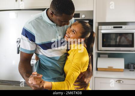 Happy diverse couple having fun slow dancing and smiling at each other in kitchen Stock Photo