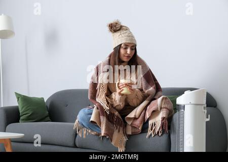 Frozen young woman with cup of tea warming near electric heater at home Stock Photo