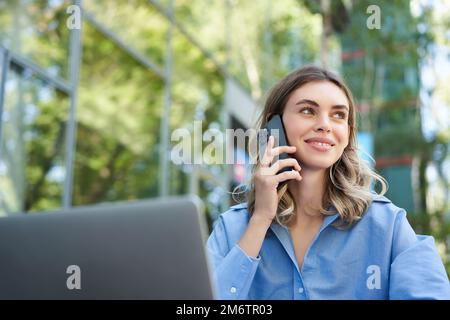 Portrait of young businesswoman sitting outside in park, talking on mobile phone, using laptop. Young woman, student working out Stock Photo