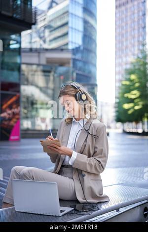 Vertical shot of smiling adult woman studying outdoors, listening to online course, taking notes, attend team meeting via app in Stock Photo