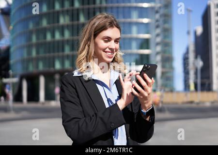 Portrait of confident corporate woman using mobile phone on city street. Businesswoman messaging on smartphone app while standin Stock Photo