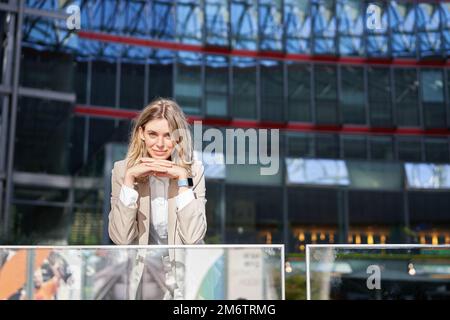 Beautiful young woman in beige suit, standing near office buildings in city center, smiling and looking dreamy Stock Photo