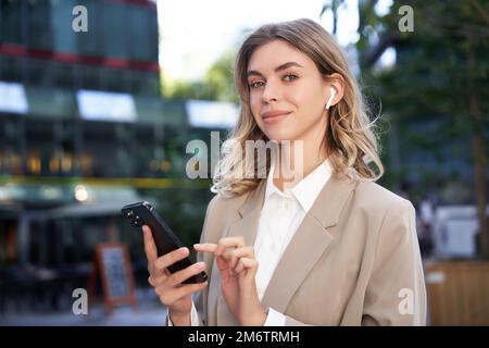 Smiling beautiful corporate woman in beige suit, standing on street in city with wireless headphones, holding smartphone Stock Photo