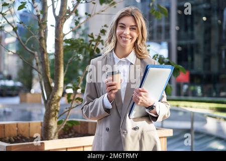 Young businesswoman standing on street with takeaway coffee, laptop and work documents, smiling Stock Photo