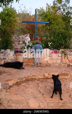 A woman kneeling and praying at the Blue Cross in Medjugorje. Free-ranging dogs pictured are a common sight. Stock Photo