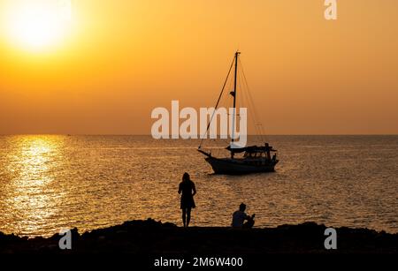 Silhouette of young couple enjoying beautiful sunset at the beach. Lonely boat moored in sea Stock Photo