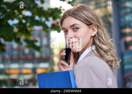 Close up portrait of corporate woman calling on mobile phone, walking on street with work documents in hand, wearing business su Stock Photo