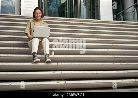 Asian girl student sits on stairs near campus, types on laptop, does her homework outdoors Stock Photo