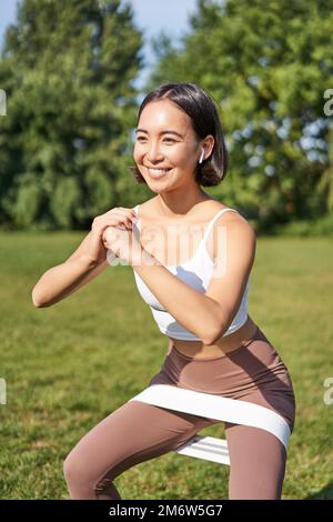 Vertical shot of young fit woman does squats in park, using stretching band on legs, smiling pleased while workout Stock Photo