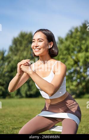 Asian fitness girl doing squats in park, using resistance band, stretching yoga rope for workout training on fresh air Stock Photo