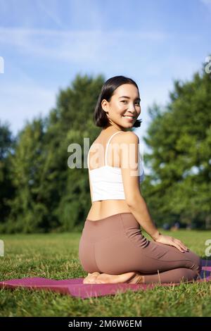 Portrait of asian woman sitting and meditating in park, doing yoga on fresh air, fitness exercises, smiling at camera Stock Photo