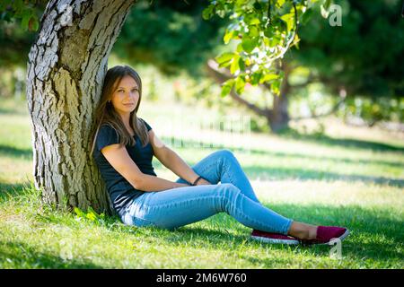 A girl sits under a tree in a sunny park and looks into the frame Stock Photo