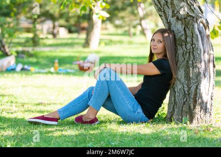 A girl sits under a tree in a sunny park and joyfully looks into the frame Stock Photo