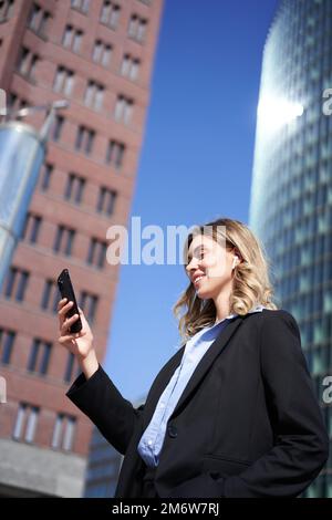 Vertical shot of successful businesswoman in suit, holding smartphone, using mobile phone on her way to work, standing with tele Stock Photo