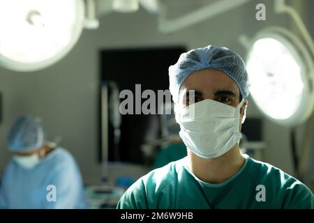 Portrait of biracial male surgeon in surgical cap and mask in operating ...