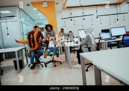 Team building and office fun. Young cheerful businesspeople in smart casual wear having fun while racing on office chairs and sm Stock Photo