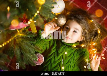 A girl with long hair and garlands lies on a red plaid under a Christmas tree with toys in a warm kn Stock Photo