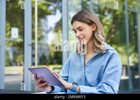 Young digital nomad, woman with tablet, working outdoors on fresh air, smiling and looking away at passerby Stock Photo