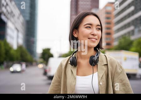 Portrait of young asian woman in headphones, posing in city, smiling and looking away, standing on street of city centre Stock Photo