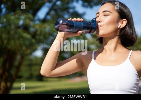 Fit asian sportswoman drinks water while running marathon. Fitness girl jogging in park, staying hydrated. Stock Photo