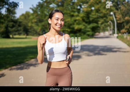Happy smiling asian woman jogging in park. Healthy young female runner doing workout outdoors, running on streets Stock Photo