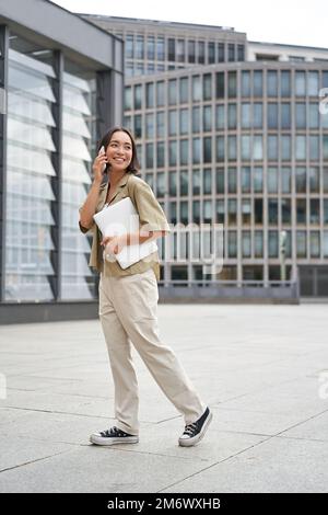 Vertical portrait of stylish asian girl student, talking on mobile phone while walking, holding laptop Stock Photo