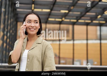Happy asian woman talking on mobile phone, standing on bus stop in city. Outdoor shot Stock Photo