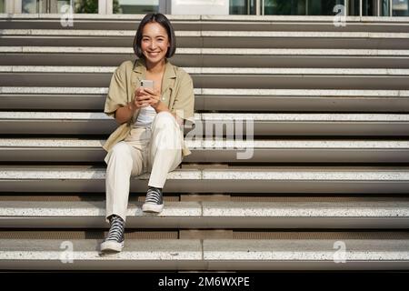 Portrait of smiling asian girl sits on stairs with her smartphone, browsing internet on mobile phone, resting outdoors in city c Stock Photo