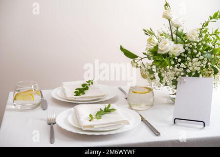 Mockup white blank space card for table number or menu. Wedding teble decoration with white flowers, glasses and white napkins. Stock Photo