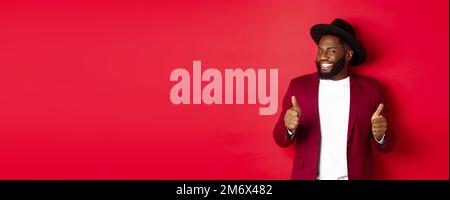 Cheerful Black man having fun on party, showing thumbs up in approval, smiling and liking something, standing against red backgr Stock Photo