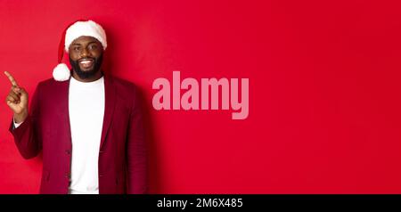 Christmas, party and holidays concept. Handsome Black man in santa hat pointing finger left, showing advertisement, standing hap Stock Photo